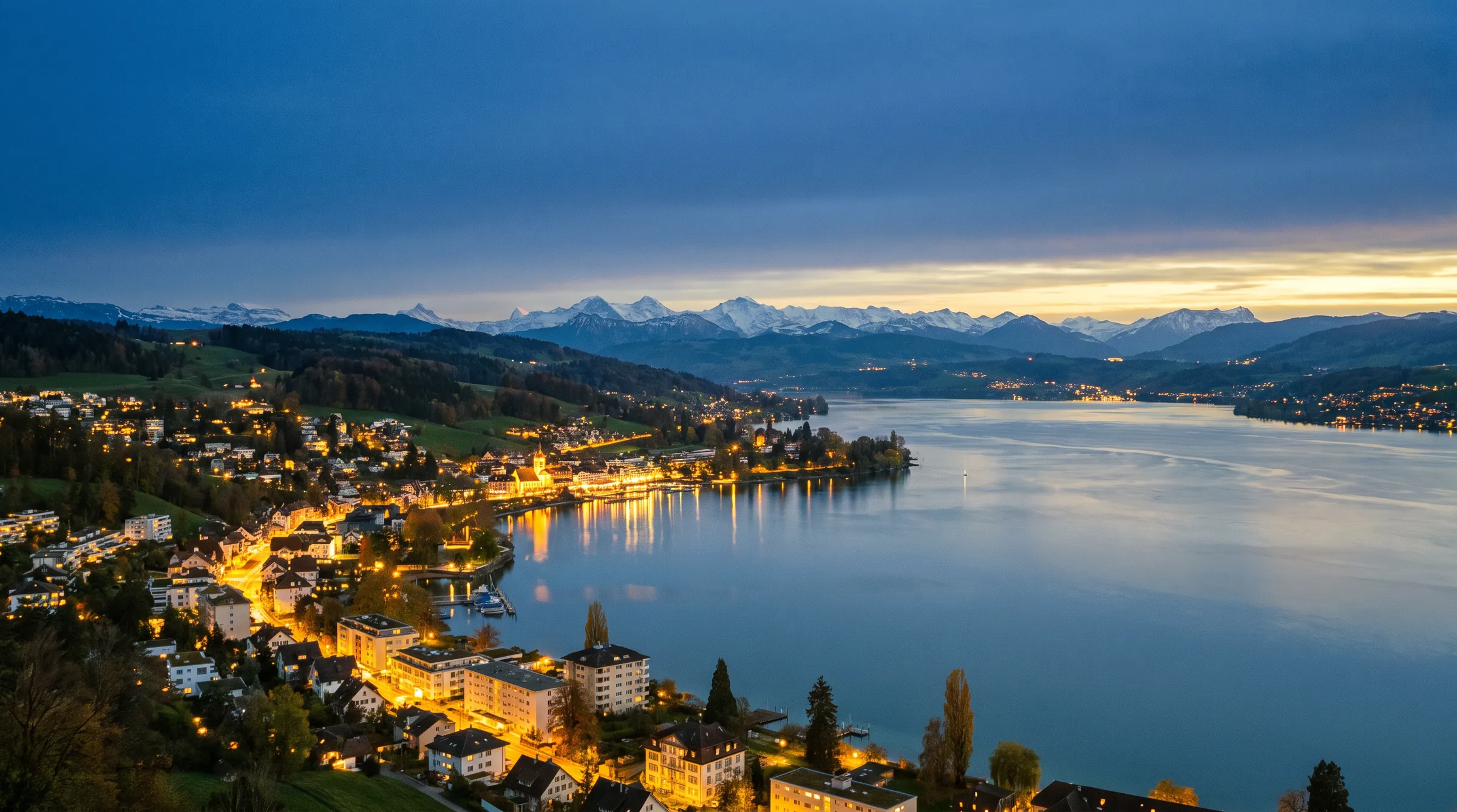 Abendstimmung über einem Schweizer See mit den Lichtern der Uferorte und den Alpen am Horizont